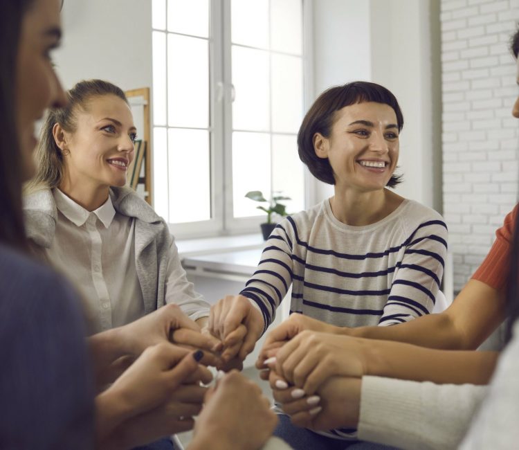 Happy smiling women sitting in circle in community meeting or group therapy session with coach, holding hands and thanking each other for support, sincere, true friendship and achieving goals together at Creo Recovery Intensive Outpatient Program